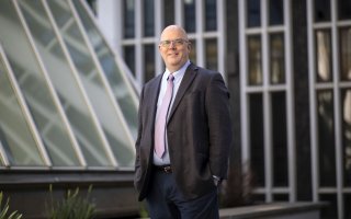 A man in a suit jacket and tie stands near glass windows