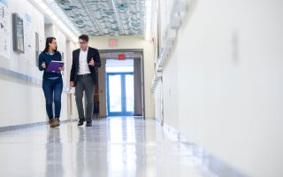 A woman wearing glassesholding a binder and pen walks forward with a man wearing glasses and a jacket in a hallway.