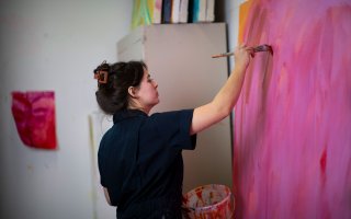 A woman in a navy jumpsuit has an arm extended as she swipes pink paint onto a large canvas frame covered in pink, orange and red paint. 