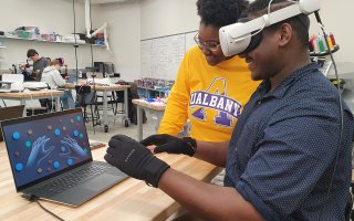 One student standing in a yellow UAlbany shirt watches another student mirror movements from gloves on a laptop screen indoors.