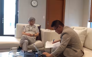 An older man with silver hair is seated in a chair next to a younger man with black hair and glasses, who sits on a couch nearby consulting a handful of papers and looking at a laptop. They are gathered around a table with a pitcher of water and other beverages.