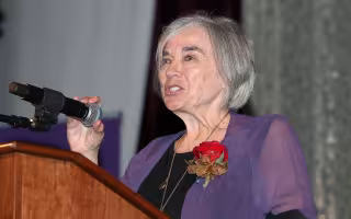 A woman with short gray hair wears a purple blouse and rose lapel as she stands at a podium with a hand grasping a microphone