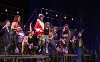 More than a dozen students dressed in 1980s and 1990s attire stand on a table on a dark stage and sing, a string of lights hang behind them. A woman in a Santa coat stands at the center singing.
