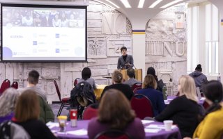 A woman speaks from a podium near a screen with a projected agenda on it. The backs of heads of the audience, seated at round tables, is seen in the foreground.
