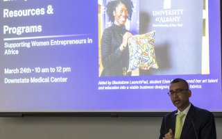 A man in a green tie speaks at a lectern with a presentation screen behind him featuring Blackstone Launchpad UAlbany student Elsie Essien.