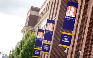 UAlbany's School of Social Welfare building on the Downtown Campus. The building is red brick, with three sets of purple and gold UAlbany flags mounted on the side. Each set of flags has the Minerva logo on top. Then below, one flag says School of Social Welfare; the other two say School of Criminal Justice.