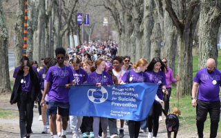 Students and community members gather to walk a course on UAlbany's Uptown Campus for the 2022 Out of the Darkness Walk. The day is sunny and a line of students leading the walking crowd hold a blue "American Foundation for Suicide Prevention" and wear purple T-shirts that say "Team Gabby". banner  