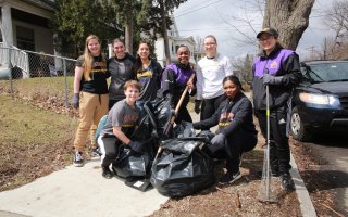 Eight people wearing UAlbany gear hold rakes and trash bags in an Albany neighborhood as they pose for a photo