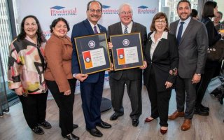 Six smiling people face the camera in a line, the two men in the center holding framed award certificates,