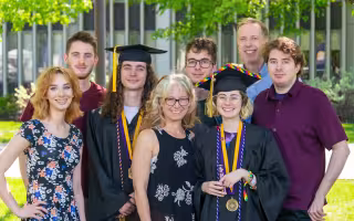 A family of six siblings and a mother and father celebrate commencement outdoors as two of the children where graduation regalia.