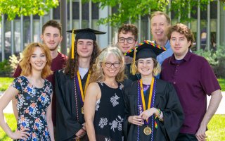 A family of six siblings and a mother and father celebrate commencement outdoors as two of the children where graduation regalia.