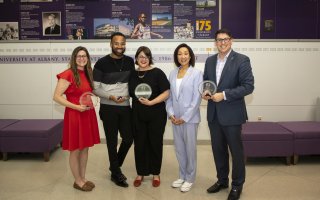 5 smiling people, four of them holding round, glass awards, stand in a line in a building atrium.