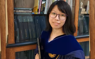 A women wearing glasses and a blue velvet PhD hood stands in front of a bookcase