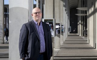Shawn Bushway stands on the pillared UAlbany Podium in a blue jacket and purple button-doown shirt