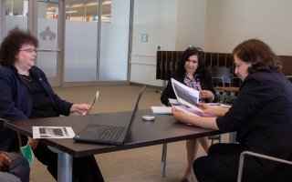 Three women sit around a table with a laptop on it, looking at printed materials