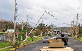 A convoy of military vehicles drive down a road in Puerto Rico with damaged power lines and light poles.