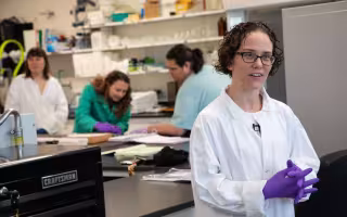 A woman with short brown curly hair and glasses wears a white lab coat and purple latex gloves, her hands clasped together as she smiles and speaks to someone outside the frame.