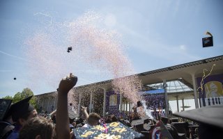 A confetti cannon shoots a blast of purple and gold confetti over a sea of black mortar board hats and hands raised in celebration at UAlbany's spring 2023 Undergraduate Commencement.