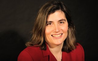 Portrait of Erin Bell; Erin is smiling wearing a red blazer with a dark brown backdrop