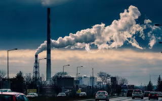 Landscape image of factory smokestacks in the background and a busy multi-lane highway in the foreground. The top layer of the sky is blue; the layer closer to the ground is cloudy with a light haze. 