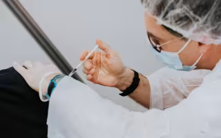 A healthcare provider wearing PPE, glasses and a blue medical mask administers a vaccine to a patient wearing a black shirt against a plain white background.