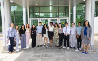 Twelve undergraduate students stand in a row in front of the Life Science building entranceway. RNA Institute director Andy Berglund is standing on the far left. Shanise Kent, assistant dean of Graduate Education, is standing on the far right. It is a sunny day, but the group is standing in the shade of an awning attached to the building entrance. Most of the building behind them is glass, with silver colored columns and trim.