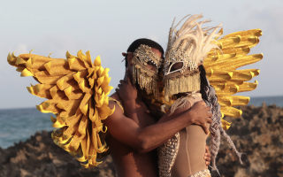 A still from the film EBBÓ shows two Afro-Dominican dancers Jeremy Antonio Caro and Rayser Rafelina Campusano Rosario embracing in elaborate costumes featuring sacred materials such as cowrie shells used in divination and rooster feathers used in rituals.