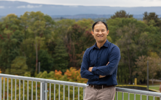 A man with short black hair in a navy shirt and tan pants leans against an outdoor railing, arms crossed, and smiles for a picture. A vast landscape of trees and hills can be seen behind him.