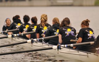 a women's rowing team is seated in a row boat with Albany Crew written on the back of their shirts.