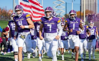 UAlbany football team runs onto the field as an American Flag waves in the background.