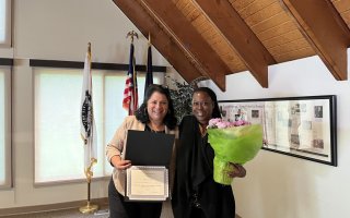 Two women stand next to each other, one holding a certificate, the other holding a pot of pale pink flowers wrapped in lime green paper. Both women are smiling. Three flags on vertical stands and a framed historical timeline are visible in the background. 