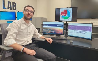 CEHC senior Ben Marzouk sits in front of a computer in the OSI Lab at ETEC with the DEA and UAlbany UCAN website homepages behind him.