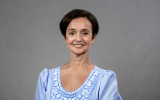 A woman with short black hairr in a bejeweled baby blue top smiles for a portrait against a gray backdrop.