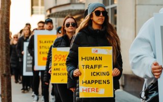 A line of people march, holding yellow anti-trafficking signs. At front is a women in sunglasses with a sign reading "Human Trafficking Happens Here"