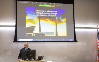 A presenter stands in front of a presentation slide during the to Pathways to Renewable Energy conference.