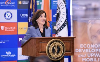 Gov. Hochul speaks from a wooden podium with the University at Albany seal, in front of a colorful poster of SUNY college logos