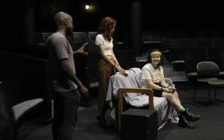 Three people appear in frame. A man stands to one side gesturing to a woman who is seated, as another woman stands directly behind her watching.