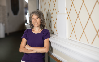 A woman with shoulder-length gray hair wears a purple t-shirt and smiles for a portrait with her arms crossed.