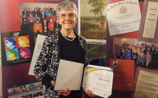 A smiling Mary McCarthy holds her award while standing in front of a panel with photos and posters 