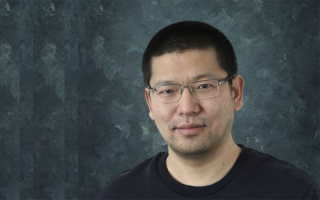 Portrait of Xin Wang, wearing a dark shirt and glasses against a green mottled backdrop. 