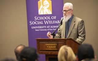 Eric Hardiman of the School of Social Welfare delivers remarks at the press event, standing at a podium that says "University at Albany". A purple banner is behind him, which reads “School of Social Welfare". Eric is wearing a tan suit jacket and glasses with black rectangular frames.