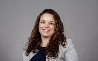 Sarah Domoff poses for a portrait against a gray background wearing a light gray jacket over a navy blue shirt. Domoff is smiling and has brown curly hair. 