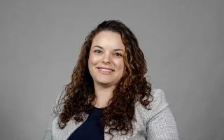 Sarah Domoff poses for a portrait against a gray background wearing a light gray jacket over a navy blue shirt. Domoff is smiling and has brown curly hair. 