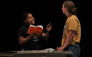 A woman holds a red book and gestures with a pen to a taller man facing her as the two rehearse for a play.