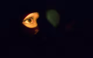 The entire image is primarily in dark shadow; the only thing illuminated is a brown human eye with dark lashes.