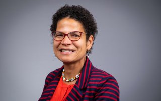 Portrait of Elizabeth Vásquez against a gray background. Vásquez is smiling, wearing red rimmed glasses, a shell necklace, red top and red and navy striped blazer.