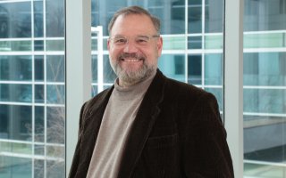 Portrait of Scott Tenenbaum, who is wearing clear-rimmed glasses, a dark brown blazer and tan turtleneck sweater. Tenenbaum is smiling, standing in front of a wall of glass windows.