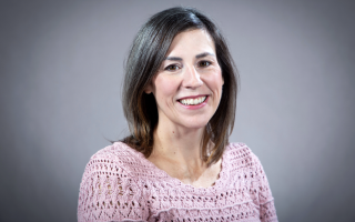 A woman with shoulder-length silver-brown wearing a pink crochet sweater smiles for a portrait against a gray backdrop.