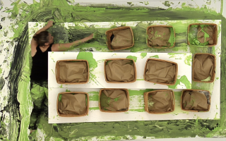 A paint-splattered woman can be seen swiping green and black paint across a canvas covered with boxes.