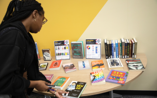 A young woman with long black braids and glasses picks up a book, titled "New Kid" off of a table topped with a diverse range of books on display.